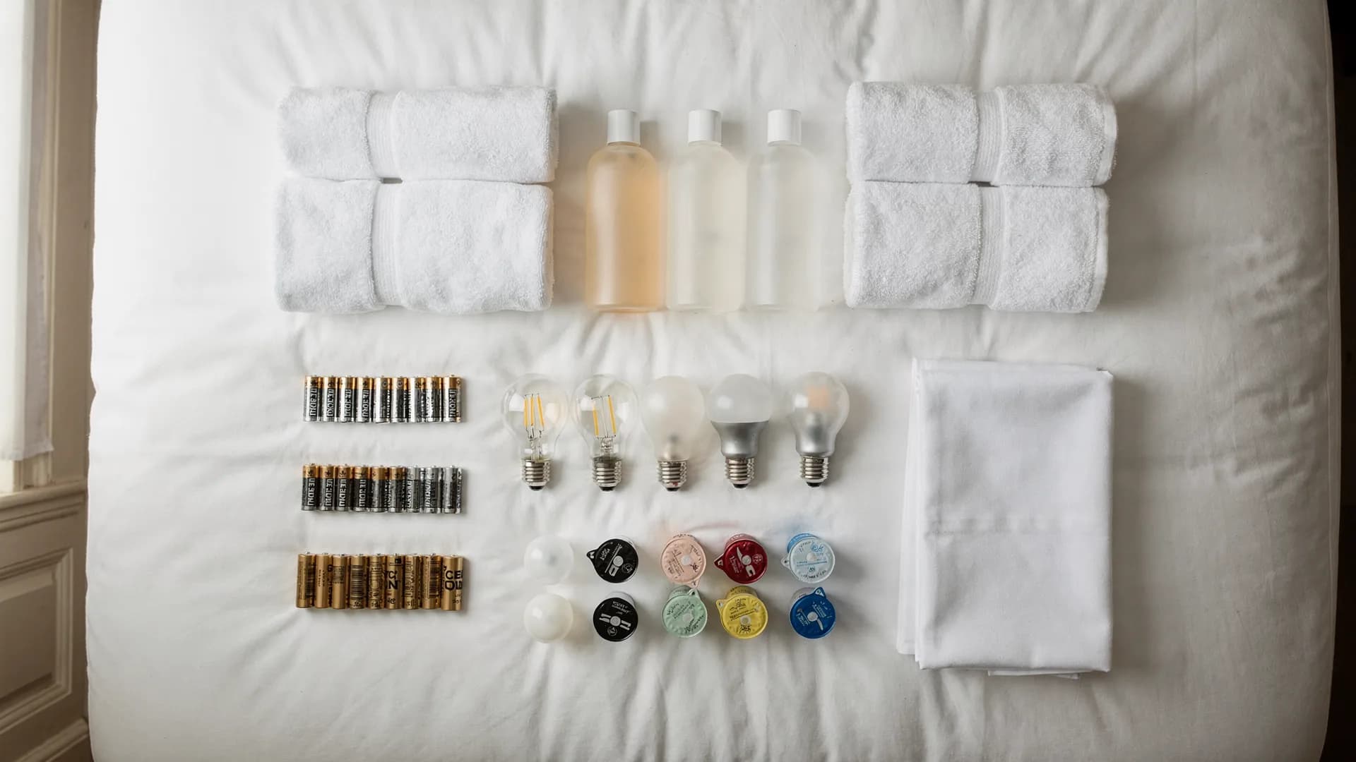Overhead view of restocking supplies arranged on a white duvet: folded towels, toiletries, lightbulbs, batteries, coffee pods, and linens.