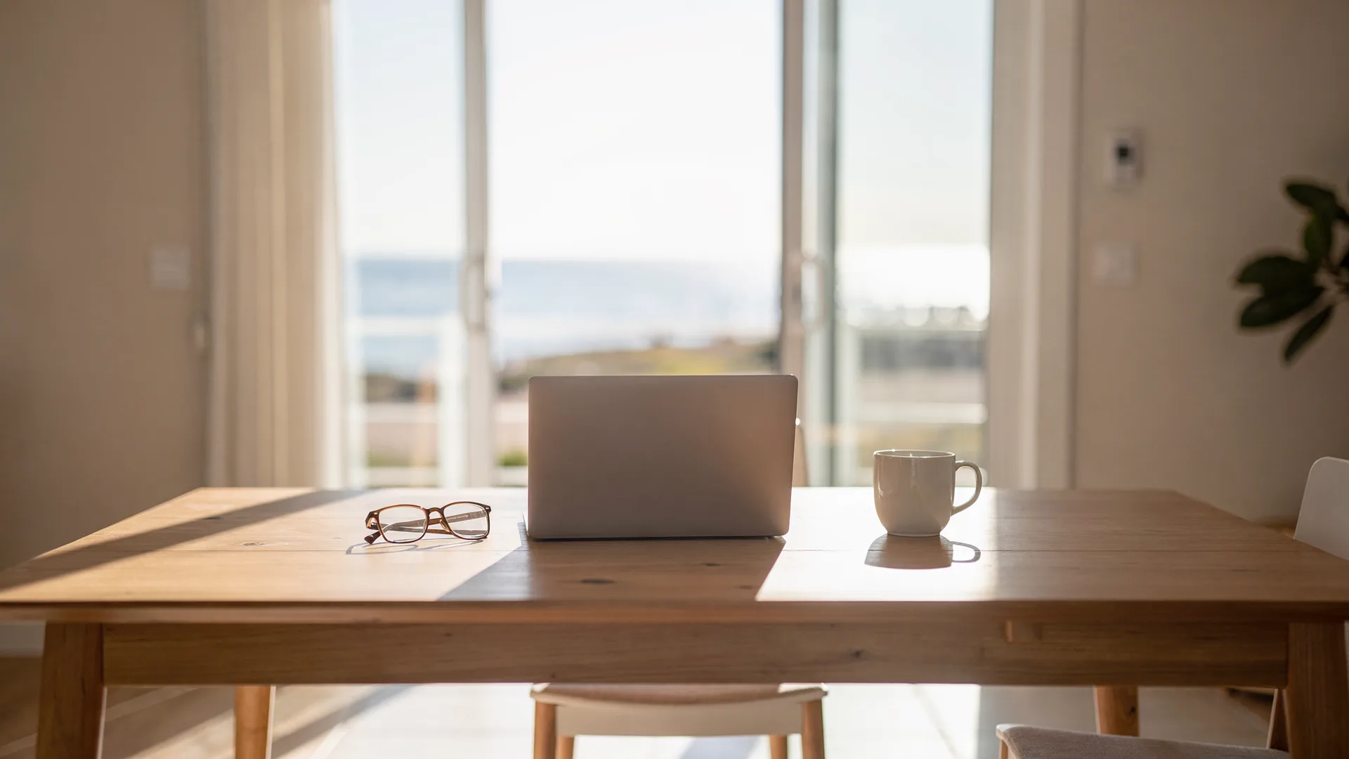 Beach house dining table with an open laptop angled away, reading glasses and a mug, coastal light through open sliding doors.