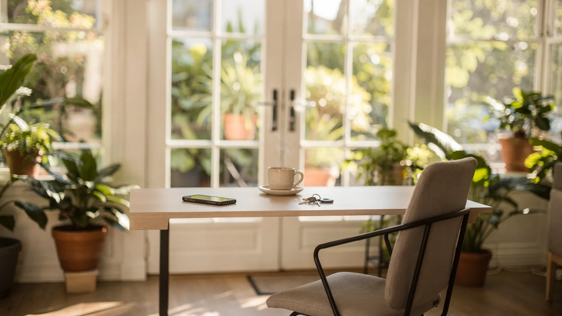 Sunroom desk in a rental with a phone lying face down beside a coffee cup and keys, chair pushed back, dappled morning light.