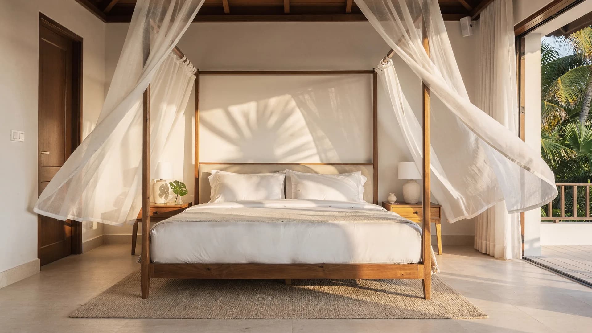 Luxurious tropical villa bedroom with a white four-poster bed and sheer curtains lifting in the breeze, flawlessly staged and empty.