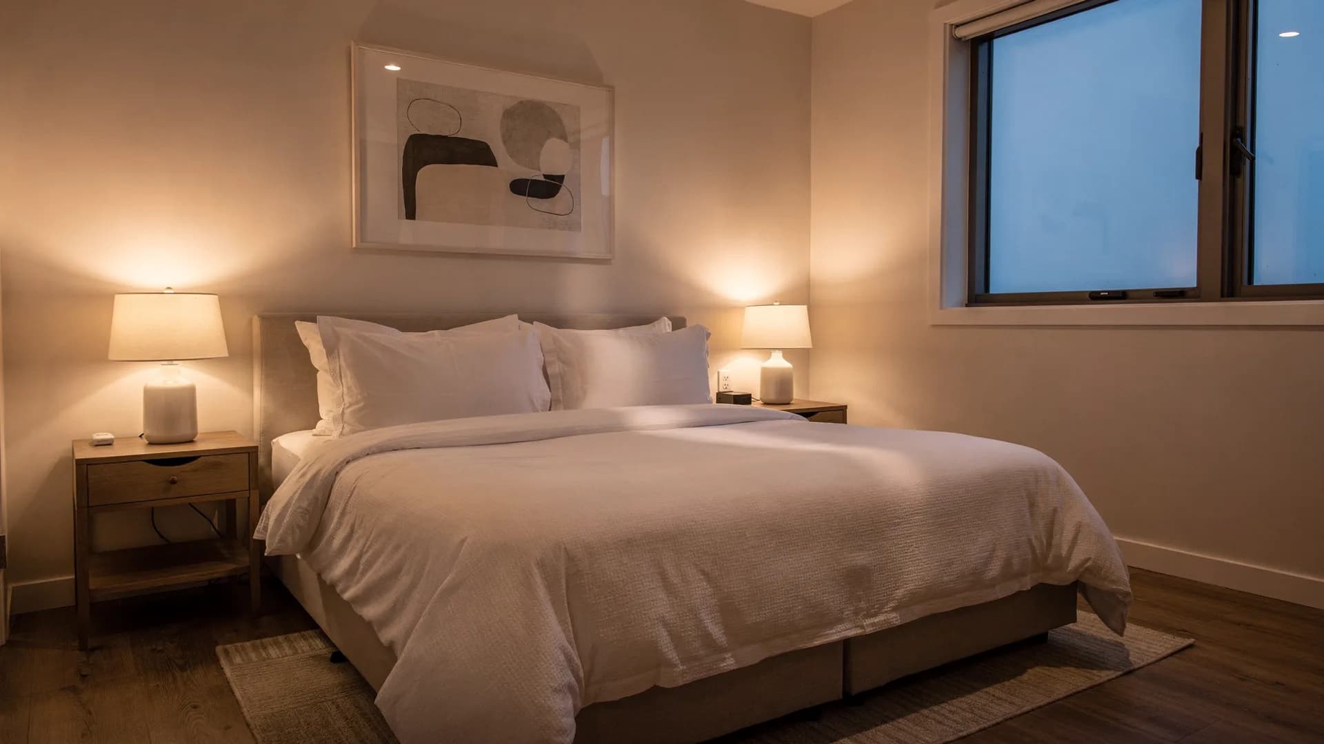 Perfectly made king bed with untouched white linens at dusk, warm bedside lamps glowing against cool evening window light.